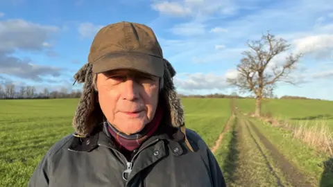 Andrew Stevenson is wearing a dark green head covering with furry flaps over the ears, a green waxed jacket and purple scarf. He is standing a few yards away from an oak tree, beside a path marked in a green field with hedges in the far distance.