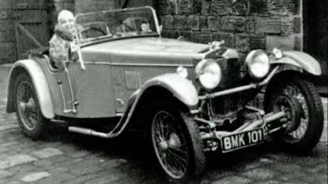 Wolf Safety Lamp Company A woman sits in an old car without a roof. The number plate on the front reads BMK101.