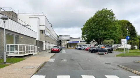 A general view of a hospital entrance. Cars are parked outside of the hospital. There are trees located on land in front of the hospital building.