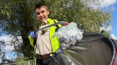 Young boy with brown hair, wearing a smart plaid shirt and yellow hi-vis smiles to camera holding a bag and litter picker.