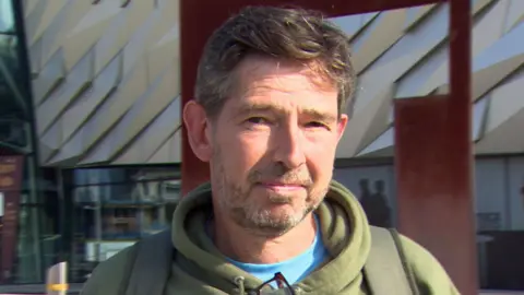 John smiling at the camera. He is wearing a green hoodie and blue tshirt. He has brown hair. The Titanic museum is behind him.