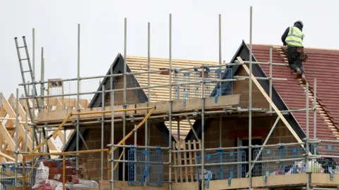 A generic shot of housebuilding, with a man tiling a roof and scaffolding against a partially built house.