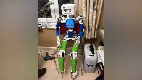 Malcolm Stern A humanoid robot prototype seated on a chair. It has a white rectangular head with two circular camera-like "eyes" giving it a simple, expressive face.
