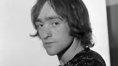 A black-and-white image of a young Dave Mason with shoulder-length dark hair. He is prominent in the photo and side on to and looking at the camera. 
