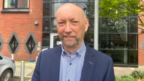 Policing Board chairman Brendan Mullan, wearing a white and blue striped shirt and navy suit. He is standing in front of a red brick building. 