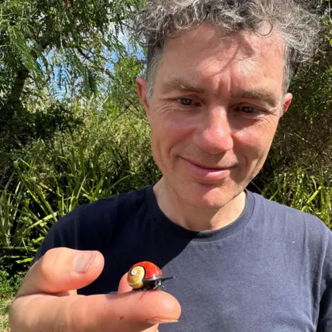 Angus Davison Smiling Man in Naval Blue T-shirt has a brightly colored snail to camera