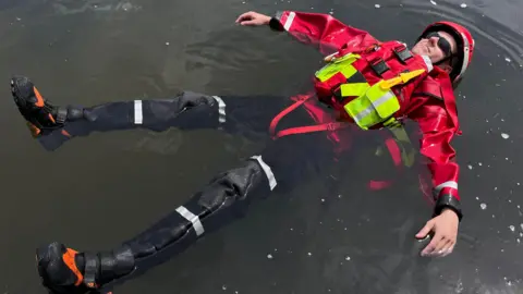 Martin Giles/BBC A man dressed in a red waterproof jacket, red helmet and black trousers and black and orange boots floating on murky-looking water.