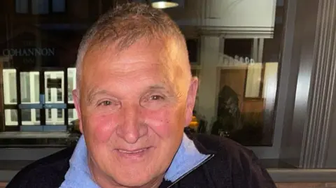 A close up image of a man with grey hair. He is wearing a navy top with a blue collar. 