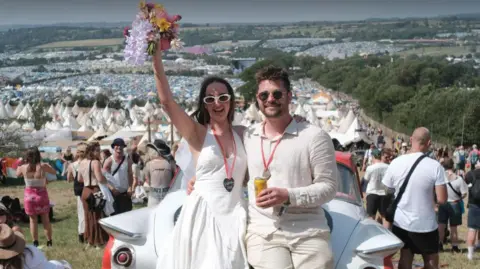 Louis Bork/Rich Brown Charlie and Charles Shires lean on a white car on a hill looking over Glastonbury Festival (2025). Charlie is holding her wedding bouquet in the air and wearing a white wedding dress; Charles is wearing a white shift and shorts and holding a yellow drinks can.
