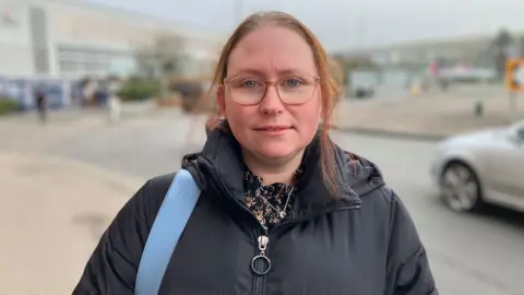 Sam Read/BBC Sandra pictured outside the Vauxhall factory which is blurred in the background. She has blue eyes and wears pale brown/pink glasses that match her hair, which is mostly scraped back. She wears a black puffa coat and has a black flowery shirt underneath, and a blue bag strap over her right shoulder. A silver car is passing by on the road.