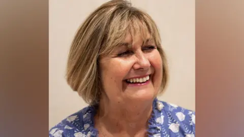 Janine Roebuck, who has a light brown blonde streaked bob with a fringe. She is smiling broadly, turning her head to the right and is wearing a blue and white cotton top 