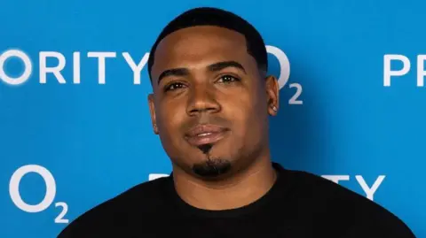 Tyrique Hyde has short black hair and a goatee. He is wearing a black crew neck jumper in front of a blue advertising board.