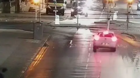 A CCTV image showing a white car with its red brake lights on as it is about to drive through some traffic lights