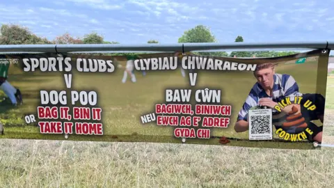 Banner in Croesyceiliog warning people against leaving dog poo. It has an image of a young boy in a blue striped shirt running with a rugby ball on it and says in England and Welsh: Sports clubs v dog poo, bag it, bin it, or take it home. 