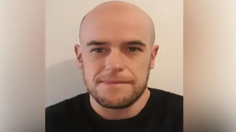 Liberal Democrats Lib Dem candidate Sean Prendergast in a black jumper positioned in front of a wall