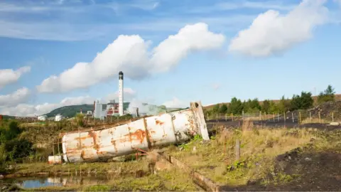 Getty Images A rusting silo on its side in a green field, with a large industrial complex in the background, with green hills behind it and blue skies above. 