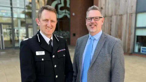 West Mercia PCC A man in a police chief constable's uniform with short light brown-grey hair stands outside, smiling at the camera. To his left is a man with short grey hair and dark-rimmed glasses, wearing a grey jacket over a pale blue shirt and a blue tie and also smiling.