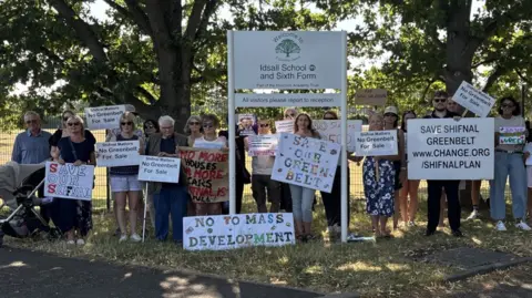 A crowd of people holding white placards with various messages opposing housing development and some large trees behind them