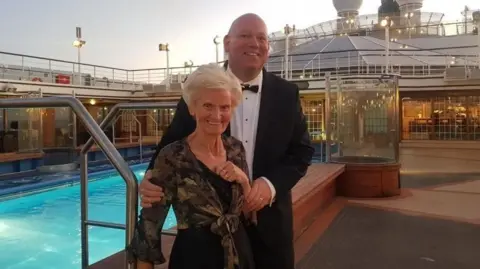 Brian Scott is standing next to his mother on a cruise ship. Behind them is a swimming pool. Brian is wearing a tuxedo and Aileen is wearing a black dress and patterned cardigan. They are both smiling at the camera. 