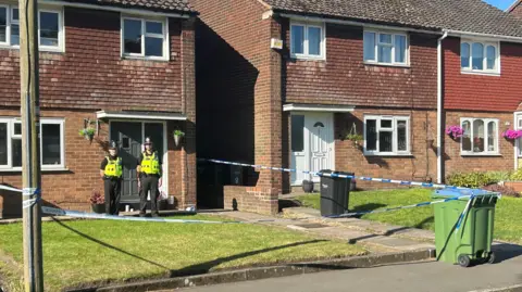 BBC Police tape around a house, with two police officers stood outside the front door