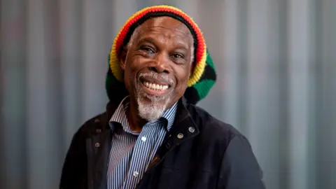 Billy Ocean is pictured smiling with a colourful head covering, striped shirt and jacket