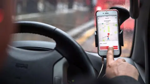 A smartphone mounted on a car dashboard displays a ride-hailing app map, with a driver’s hand reaching towards the screen, seen from inside the vehicle on a wet London street.