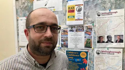James Grant, BBC Man with short dark hair, beard and glasses stands in front of a noticeboard.