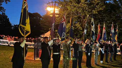 Rushmoor Borough Council A row of people, in suits and military uniforms holding large flags in front of a burning beacon. Hundreds of people are gathered behind barriers in the background.