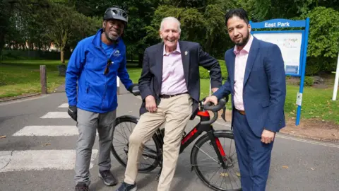 Three men standing in front of a racing bike on a path next to a park. A sign in the background reads East Park. A man at the centre with white hair in beige trousers, pink shirt and blazer leans against it, holding the handlebar. A man on his left is in a blue jacket and cycling helmet, and on his right a man in a navy suit with pink shirt. 