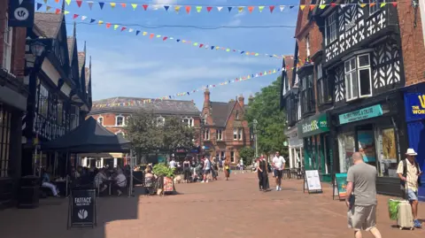 A view of Nantwich on a sunny day There are people walking down the street, as well as a number of people on the left hand side of the image sitting outside a local cafe.
