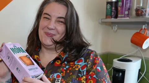 Louise Brown smiling and looking down the camera. She is standing inside a kitchen area; there is equipment on the counter behind her and an orange mug hanging from a hook under a shelf on the wall on the right of the image. There are cans of coffee on top of the shelf. Louise has straight, shoulder-length hair, that is curling around her cheeks and chin. She is wearing a heavily patterned, flowery top and is holding a carton of 'barista oat' drink.