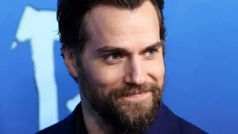 A man with grey eyes, a strong jaw and brown beard, smiles in front of a blue background.