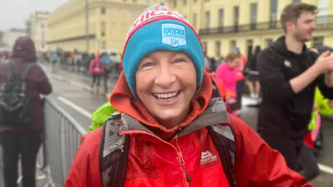Josh McLaughlin/BBC Amanda Collier stands at the barricade in the crowd near the starting line of the Brighton Half Marathon 2026. She is wearing a vibrant beanie with a logo displaying 'Chicago 5K' on the front. She beams a smile at the camera.