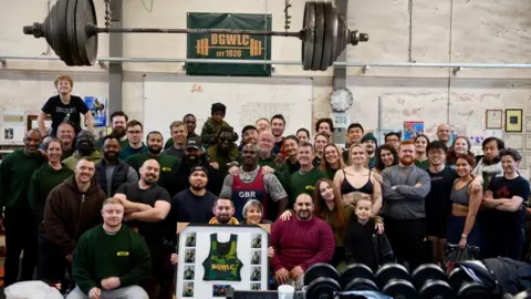 Charlotte Macaulay Two dozen people pose for a picture taken in the gym with a large weight above them