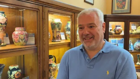 Will Moorcroft, owner of Moorcroft Pottery, is stood in front of a series of cabinets displaying Moorcroft products. He is wearing a light blue polo shirt, and has grey hair.