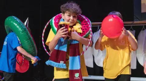 Hollis Photography Three children are standing on a stage wearing yellow and blue t-shirts. They are holding objects like hula hoops and balloons, and are wearing scarfs and rubber rings