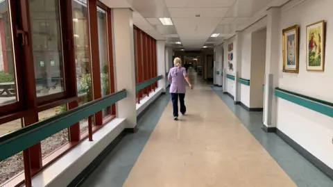 Nurse in light purple tunic and dark trousers walking down an empty hospital corridor.