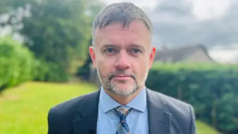 Det Insp James Callander, man with greying hair and stubble, wearing light blue shirt, blue tartan tie and blue jacket, looking at camera, with a garden in the background.