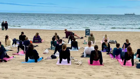 Adela Rokytova A large group of people are on the sand of a beach. Each person has their own yoga mat beneath them. These mats are various colours including blue, pink and purple. Each member of the group is holding themselves up in a bridge position, with their arms behind their back and their hands and feet on the floor. They are all facing the sea, and at the front of the group is an instructor showing them what to do.
