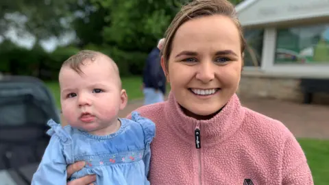 Joanne Marshall has brown hair tied back from her face and is wearing a pink fleece top. She is holding a baby in a blue dress with little pink flowers on it