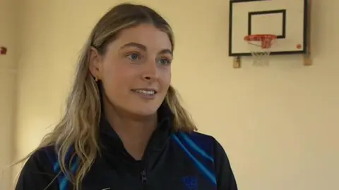 A young woman with blonde hair who is mid-sentence. She is side-on to the camera and is wearing a black sports top with electric blue diagonal lines on the shoulder. There is a basketball hoop to the top right as she is stood in a sports hall with cream walls.  