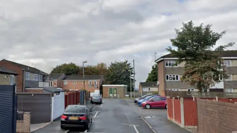 A residential street on a cloudy day. There is a row of houses on the left, and  there is a three-storey block of flats on the right. There are cars parked on both sides of the street.