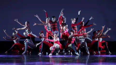 David Felton Photography A large group of dancers, all wearing red, white and black costumes, with their arms out. They are on a stage striking several poses with their arms out. 
