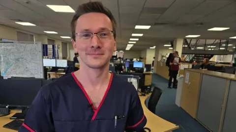 Gavin Harrison looks at the camera. He wears a navy nurses shirt with red piping. He has glasses and short dark hair. An office is visible in the background. 