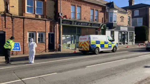 BBC A police officer wearing a high-vis yellow coat props up a sign that reads "police slow" while speaking to a person dressed in a full white forensic outfit. A white police van is parked outside a shop with police tape wrapped around the front path with orange cones marking the scene.