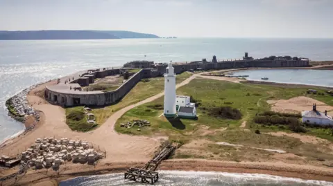 English Heritage Hurst Castle, with its perimeter spanning the tip of the shingle bank, and a white lighthouse in the foreground, pictured at the tip of Hurst Spit.