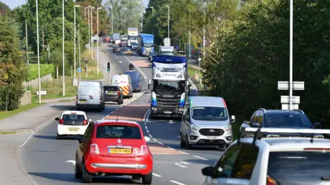 Gloucestershire County Council Traffic on the A46 in Ashchurch. It is a slightly curved road with three lanes and trees lining either side. There is a mix of vehicles including cars, vans, and lorries. 