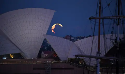 Getty Images A pink supermoon obscured by clouds seen alongside the Sydney Opera House in Australia