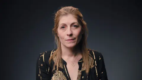 A woman with long light-brown reddish hair is looking straight ahead. She is wearing a black top, with golden threads. She is wearing a Star of David, set in a heart, around her neck