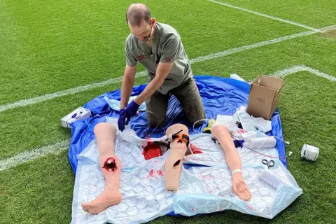 Barney Green, a consultant surgeon at James Cook University Hospital in Middlesbrough, is sitting on his knees on a waterproof floor-covering on a football pitch. In front of him are artificial limbs with fake knife injuries and fake blood, which with he is demonstrating how knife injuries look and how to treat them.